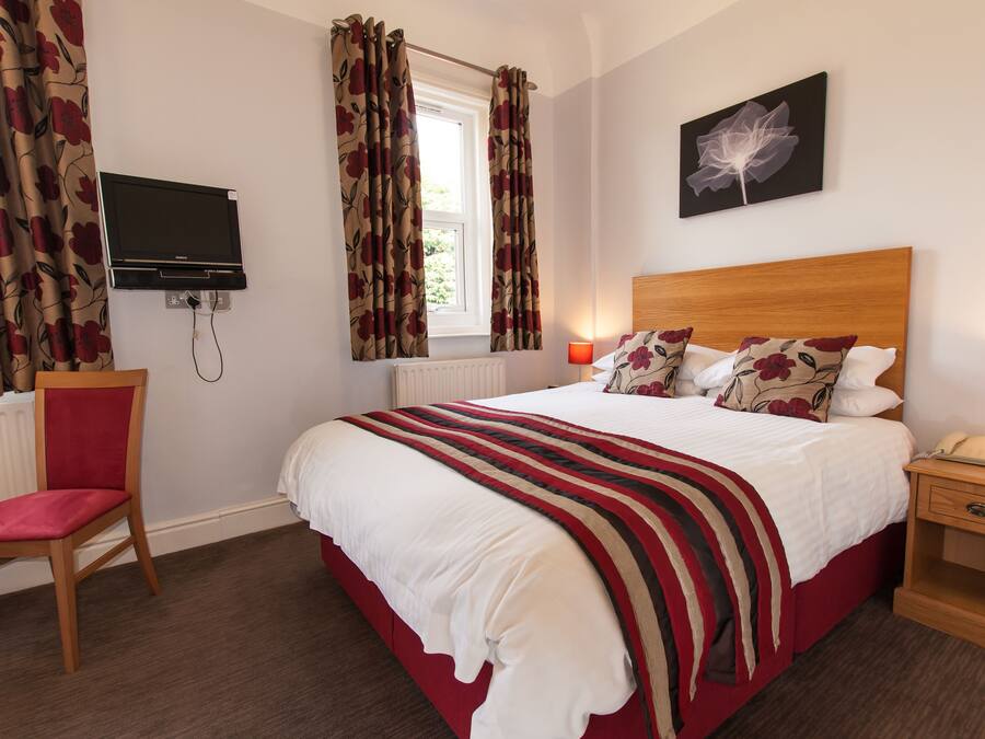 A hotel room with a bed, a chair, a TV, a window with curtains, a bedside table with a lamp, and a wall-mounted artwork.