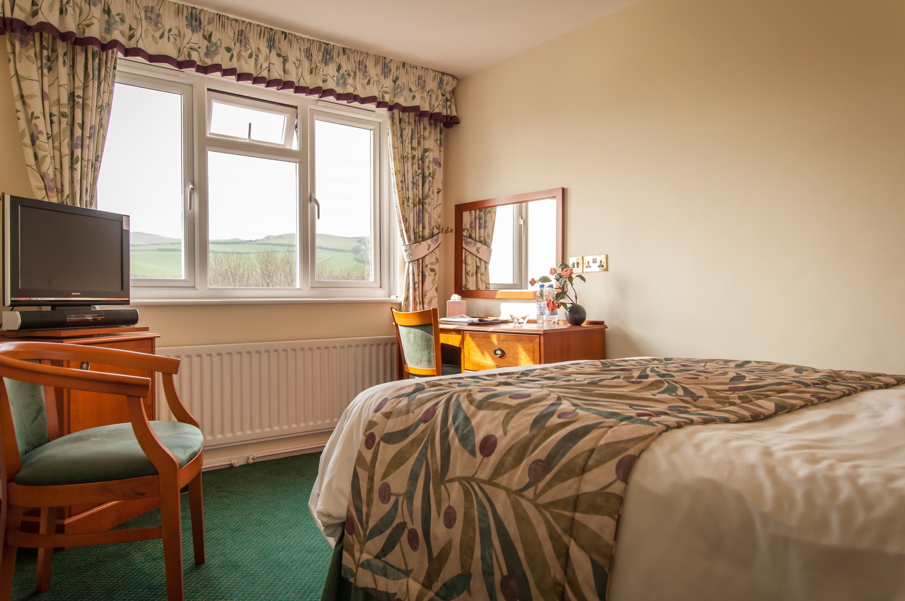 A hotel room with a bed, a desk with a computer, a chair, a window with curtains, and a radiator.
