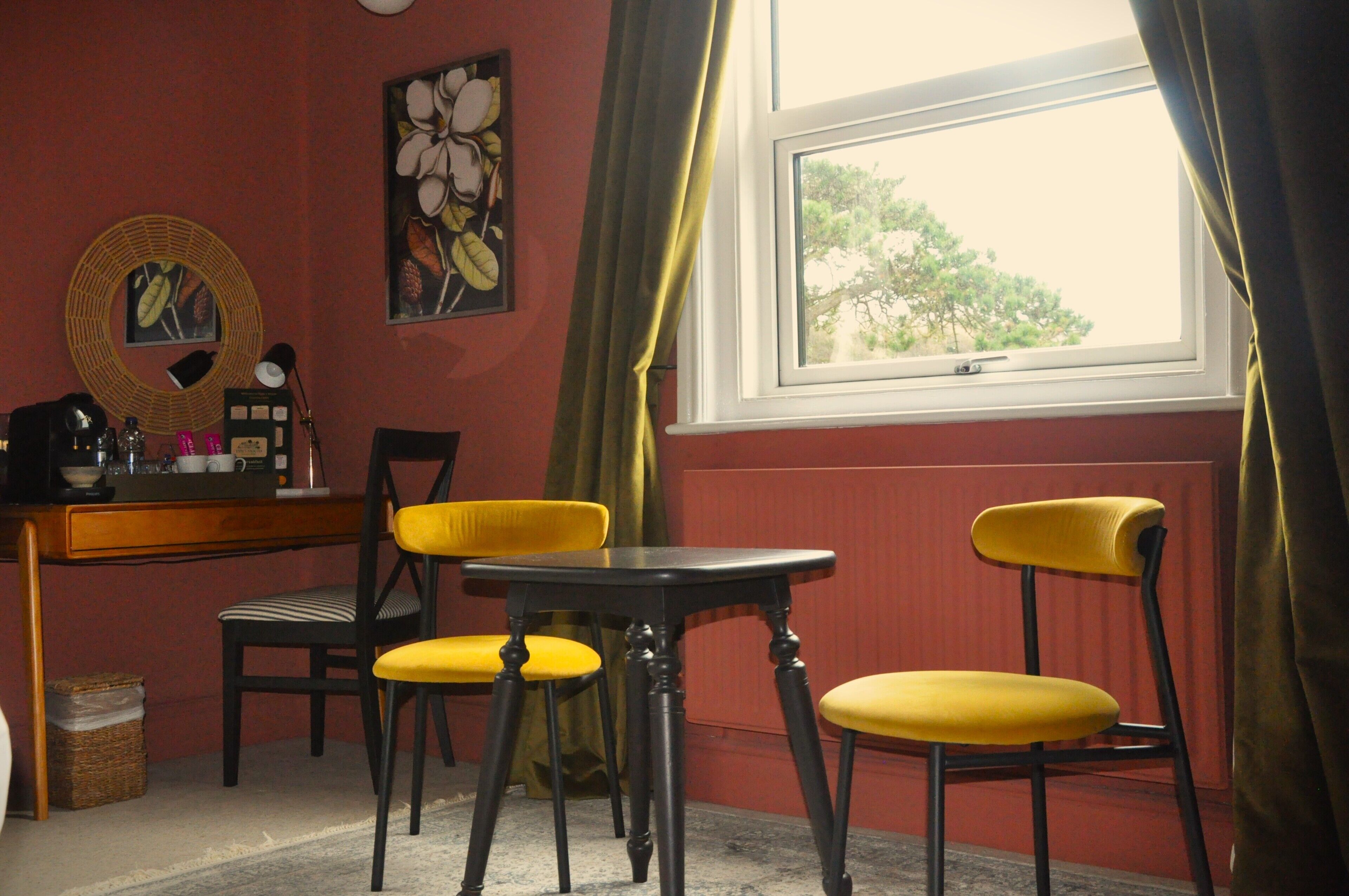 A room with a desk, two yellow chairs, a small table, and a window with curtains.
