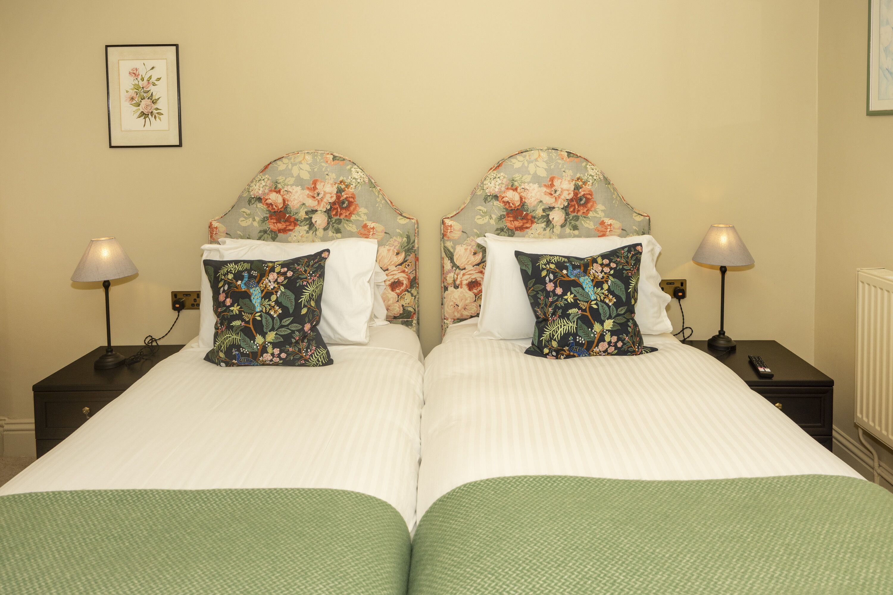 A double bed with floral headboards, two bedside tables with lamps, and a framed floral artwork on the wall.