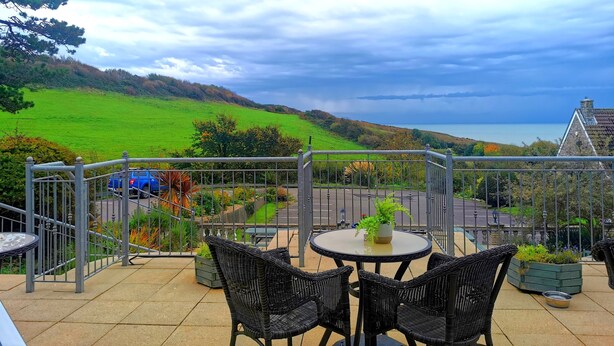 A terrace with wicker furniture and a table, overlooking a green landscape and the sea under a cloudy sky. - Eype's Mouth Country Hotel (Bridport)