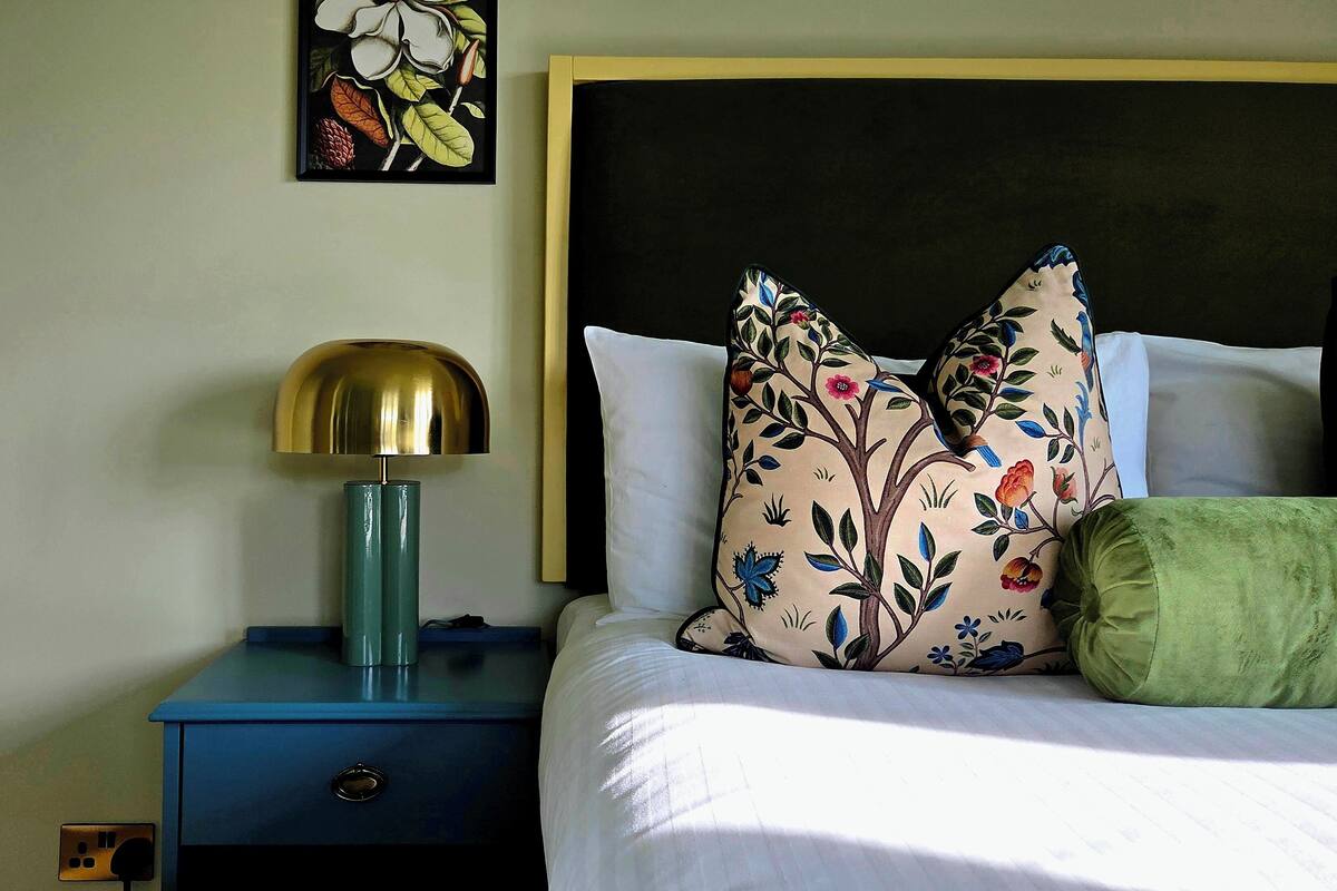 A neatly made bed with floral pillows, a green throw, and a gold lamp on a blue nightstand.