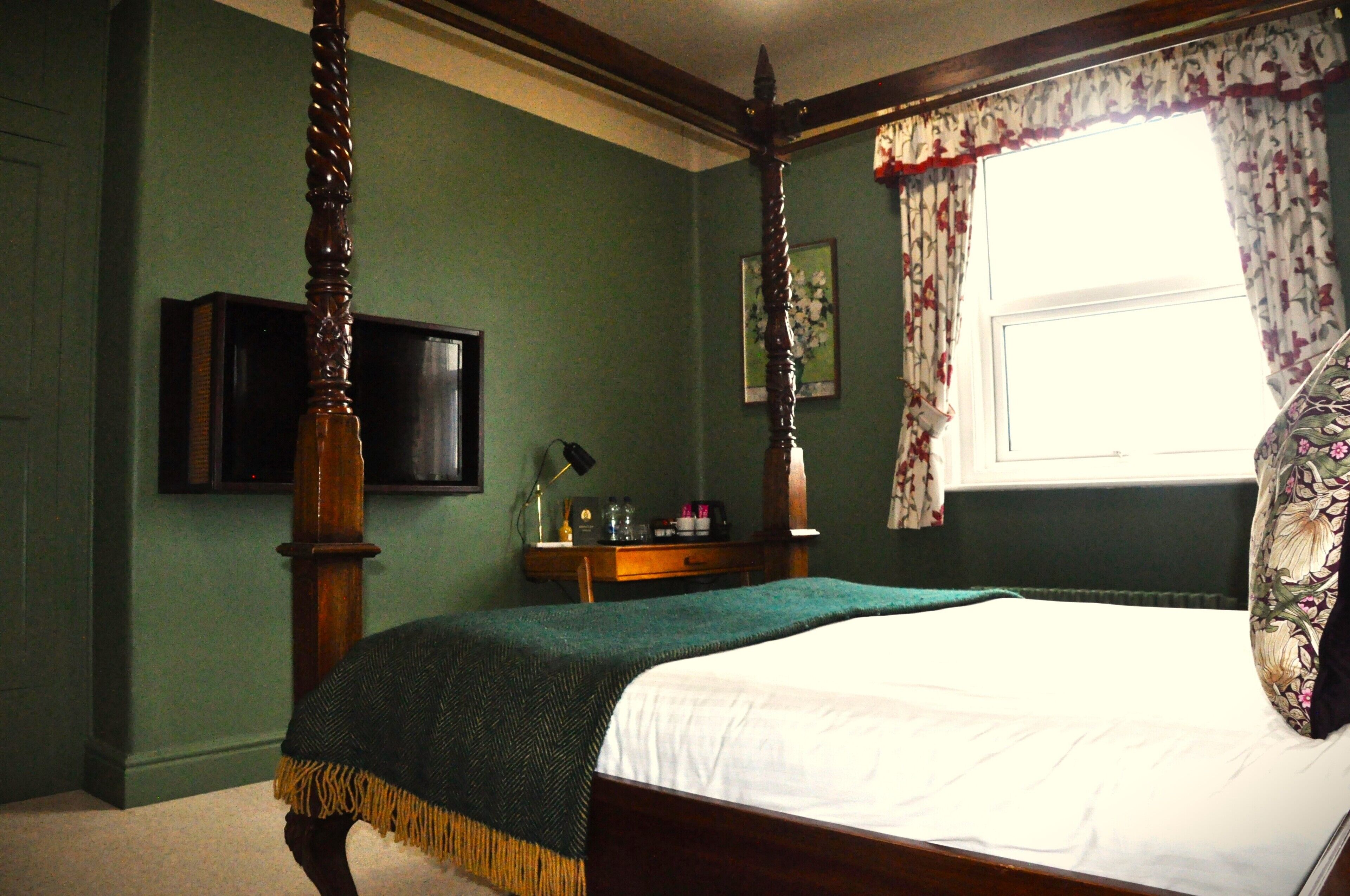 A bedroom with a four-poster bed, a television on the wall, a bedside table with a lamp, and a window with floral curtains.