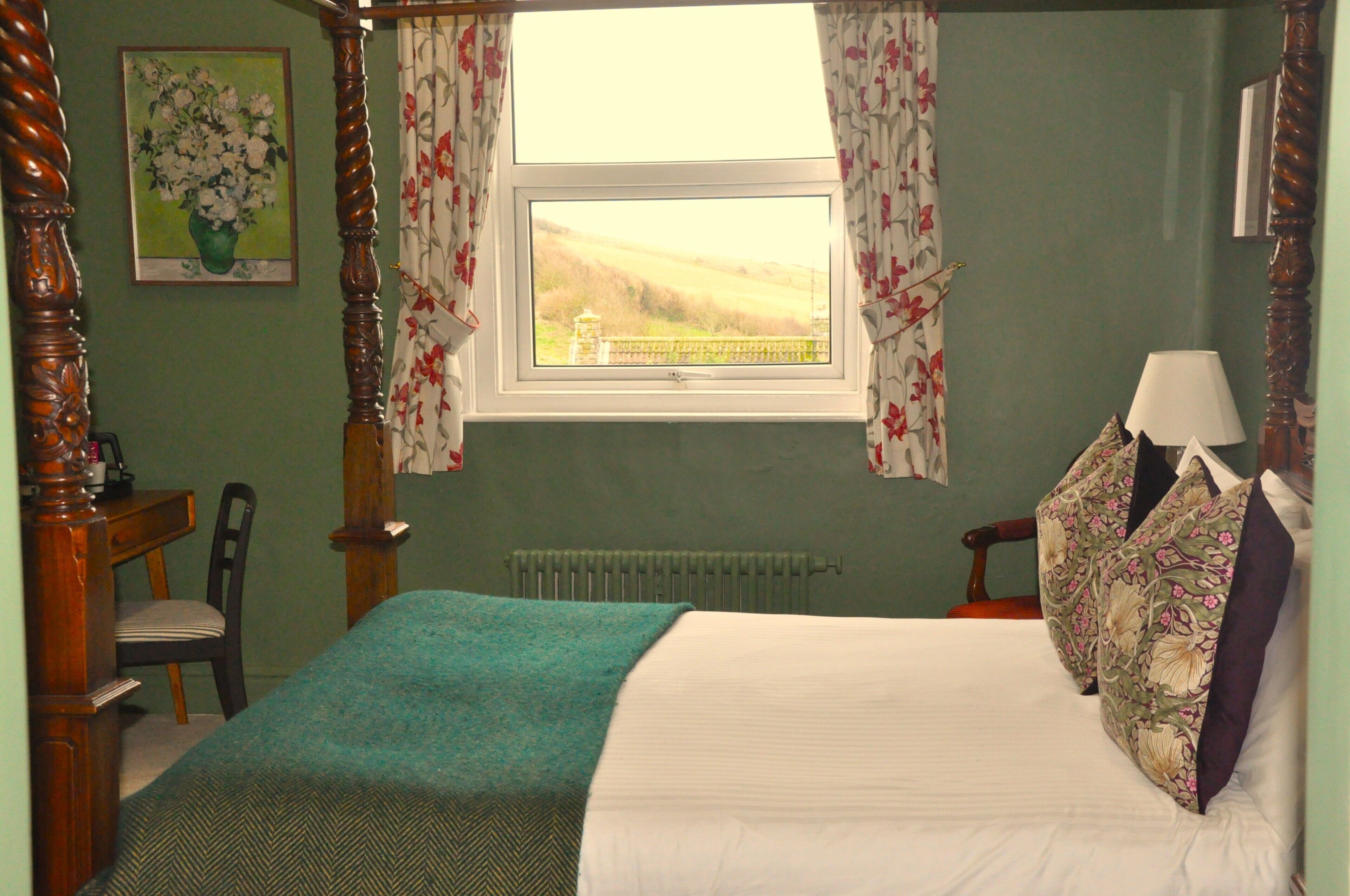 A bedroom with a four-poster bed, a window with floral curtains, a radiator, and a chair.