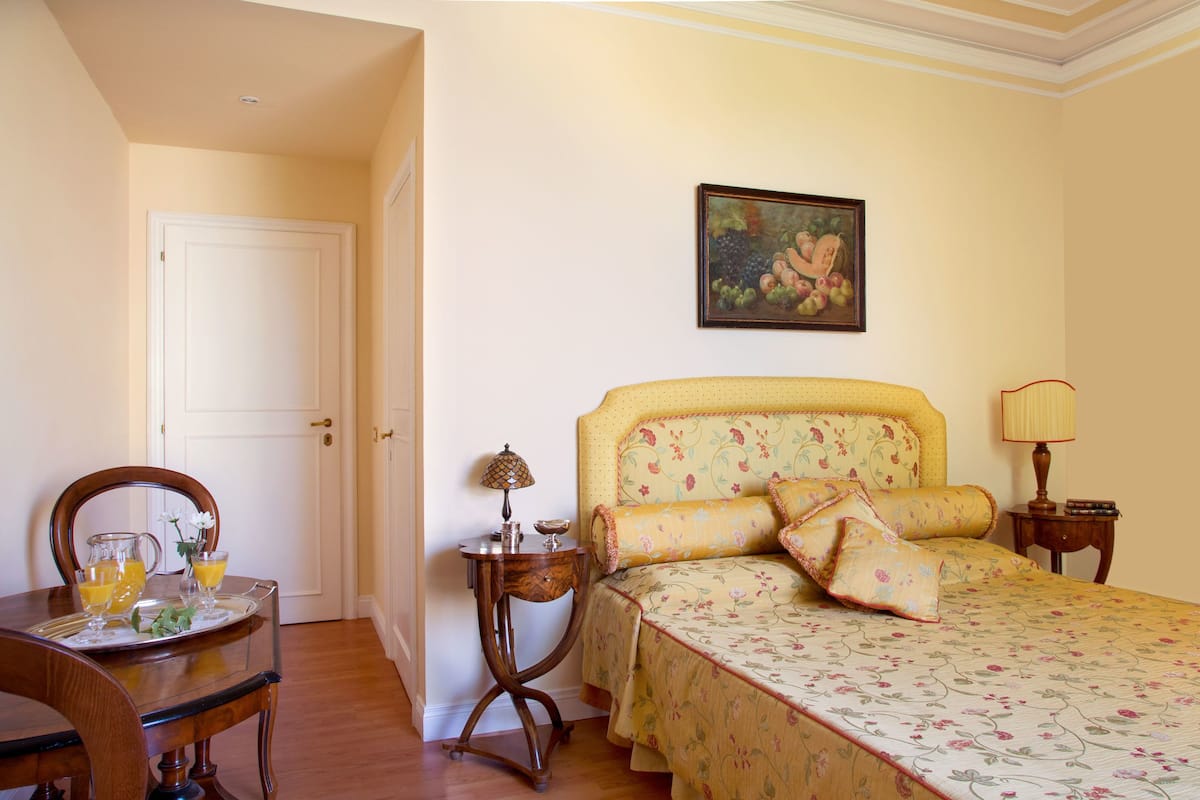 A bedroom with a bed, a bedside table with a lamp, a chair, and a table with a tray of breakfast.
