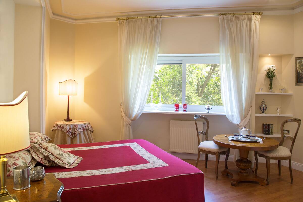 A bedroom with a bed, a bedside table with a lamp, a window with curtains, a small table with chairs, and a shelf with decorative items.