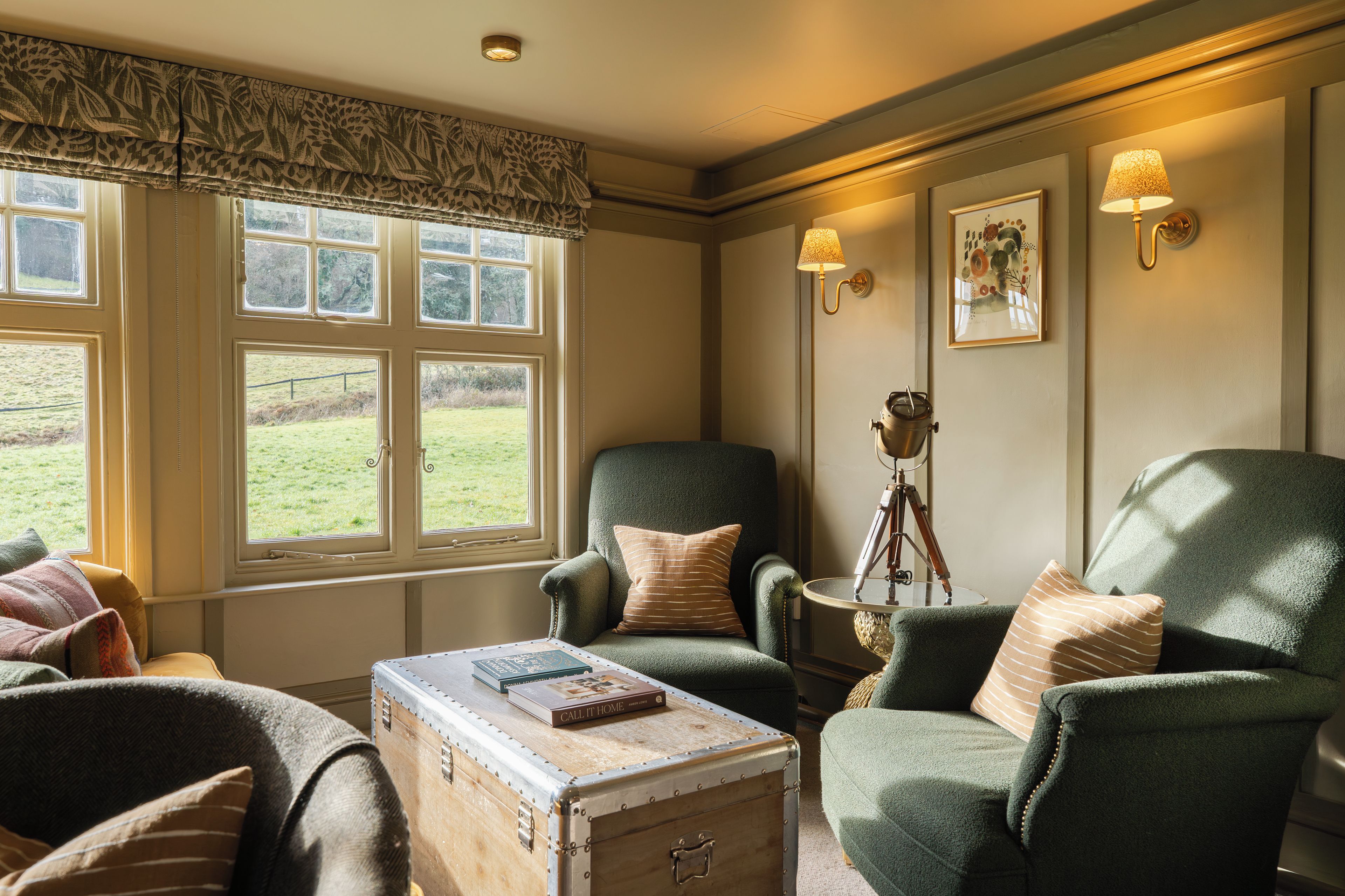 A cozy room with a large window, a green armchair, a vintage suitcase, and a tripod lamp.