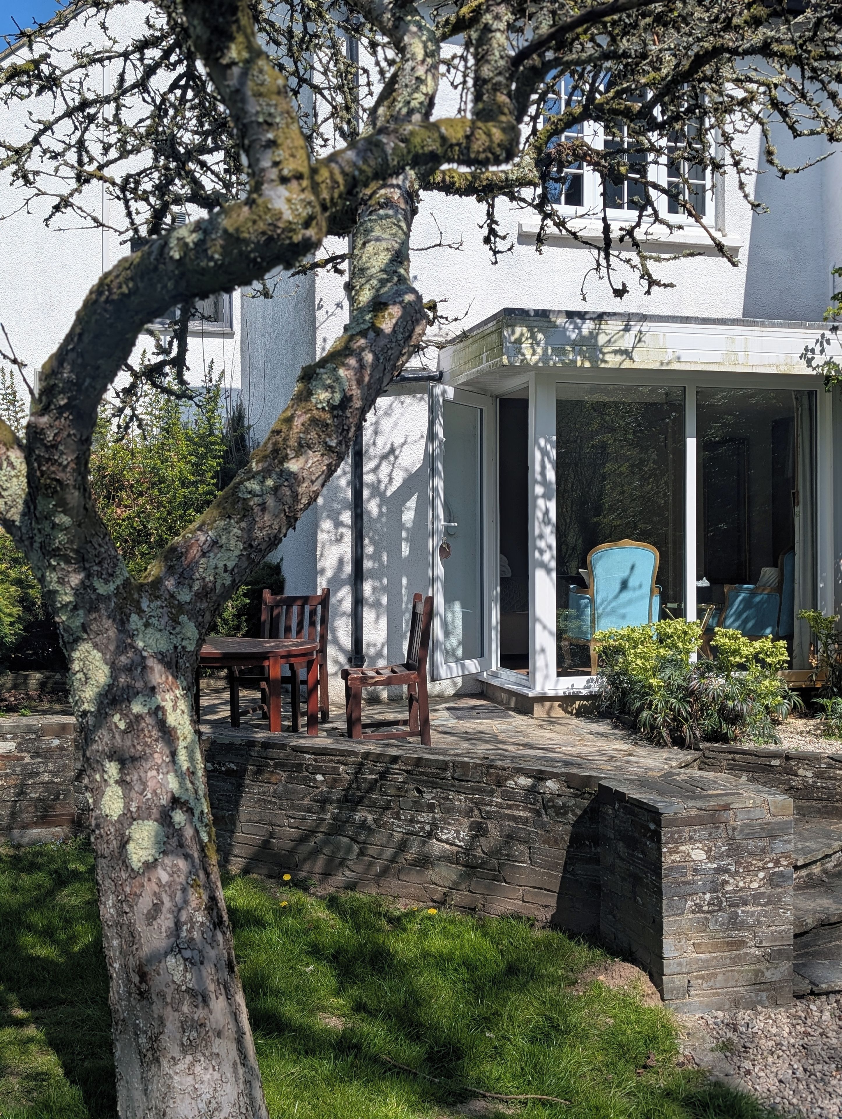 A white building with a covered patio area, a stone wall, and a tree in the foreground.