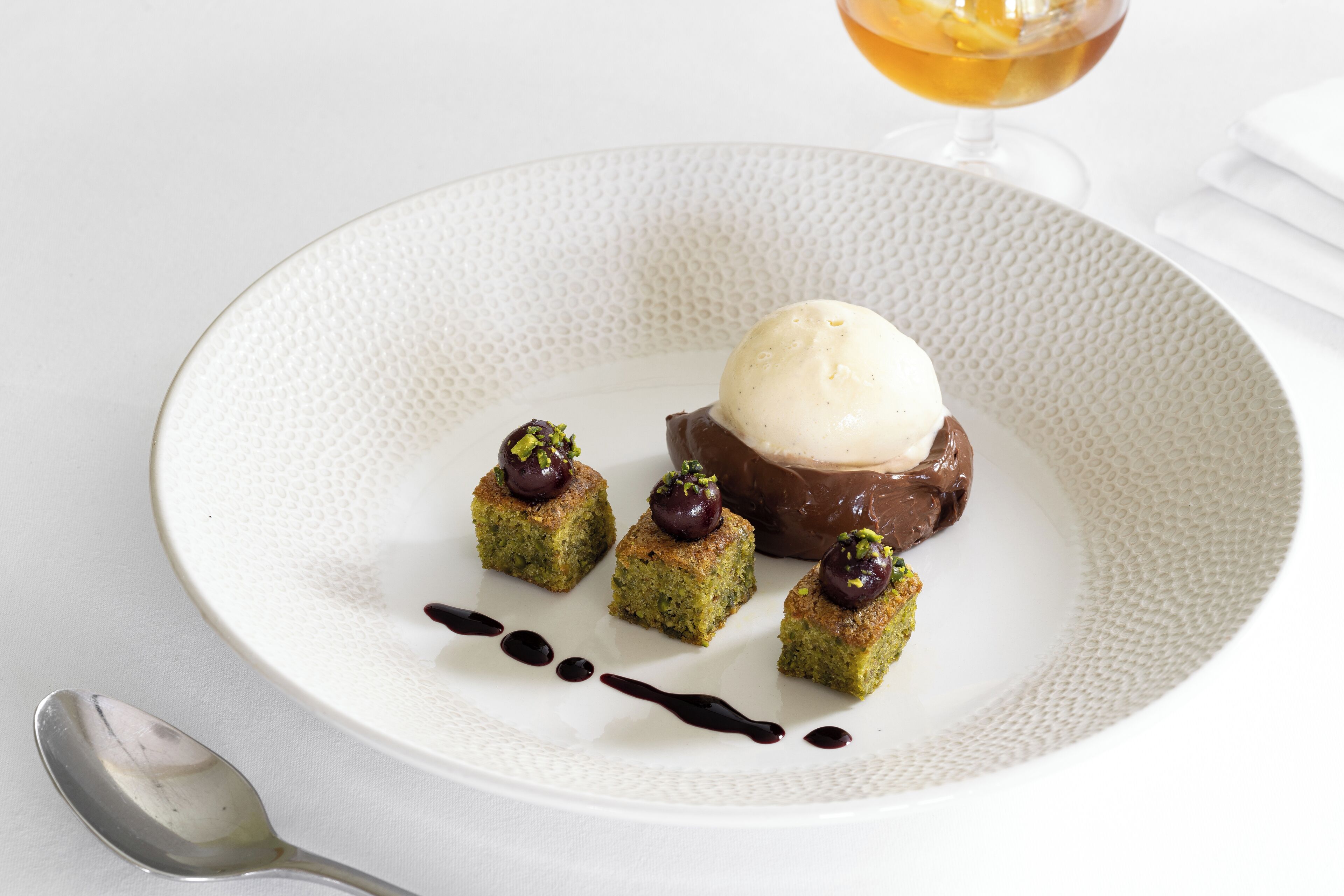 Une assiette de dessert avec une boule de glace, un dessert au chocolat et quatre petits carrés recouverts d’une cerise et de pistaches.
