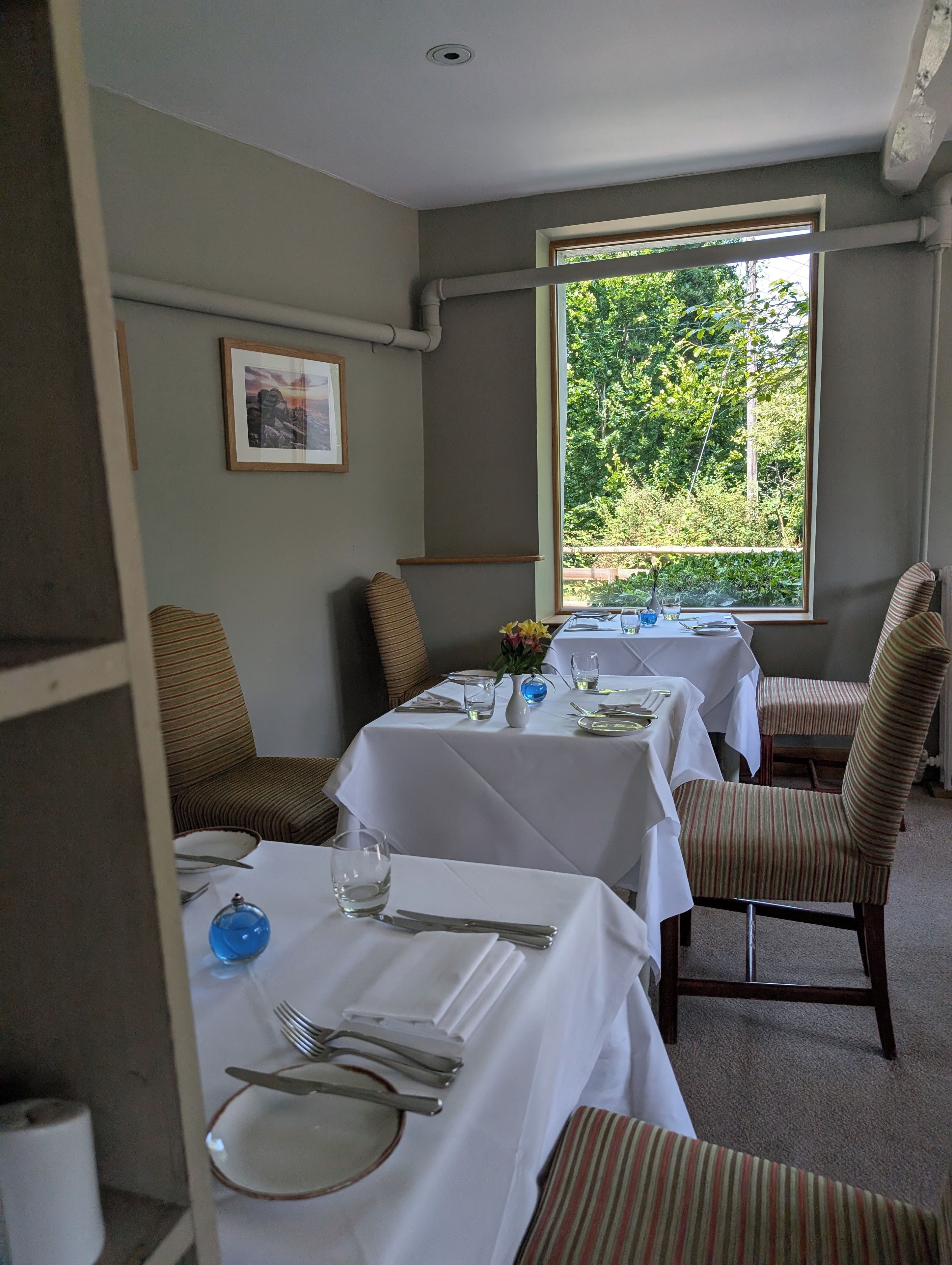 Un comedor con mesas preparadas para dos, sillas y una ventana con vistas a la vegetación.