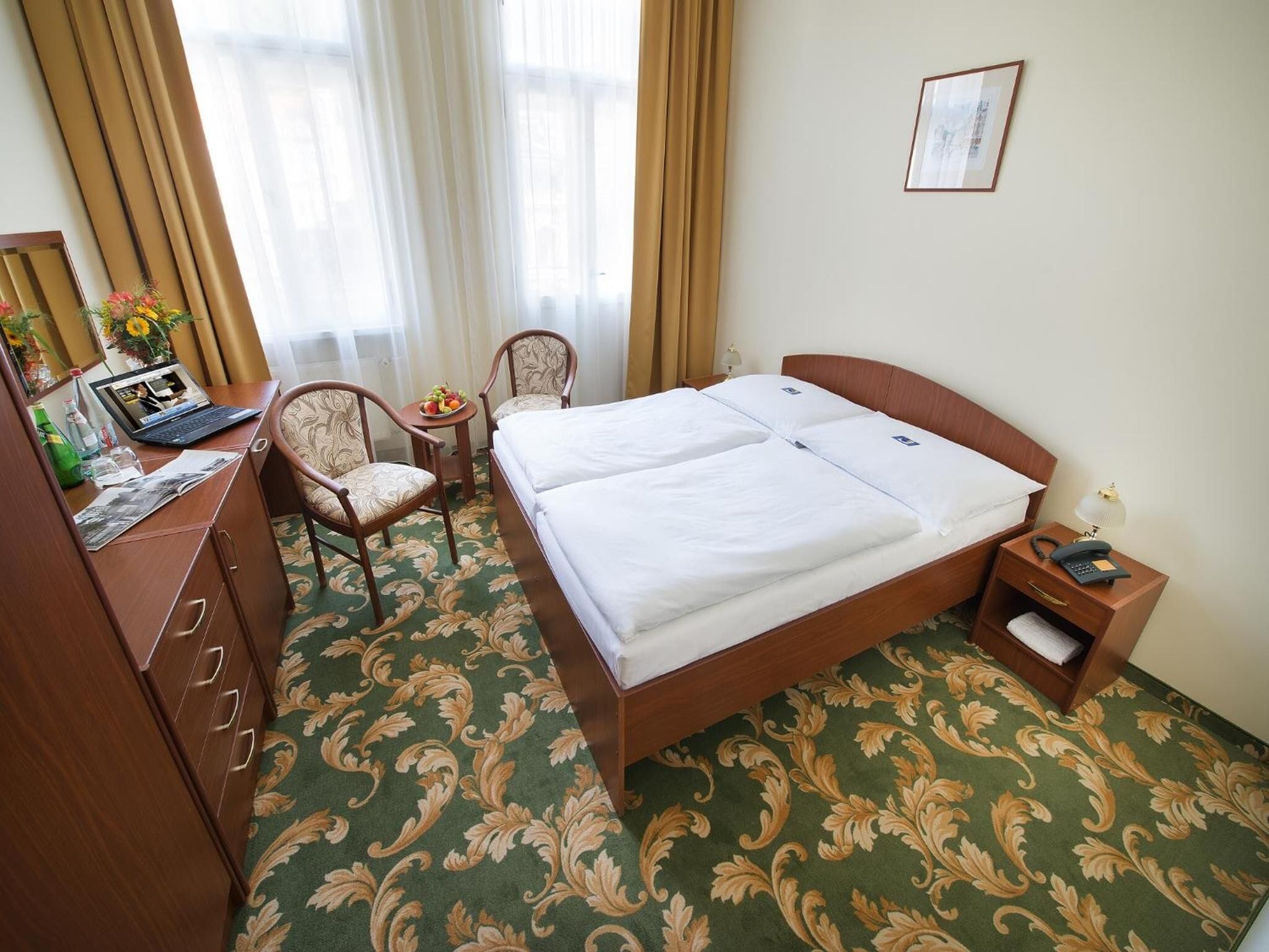 Habitación de hotel con cama doble, escritorio con una computadora portátil, dos sillas, una mesita con fruta y una mesita de noche con lámpara y teléfono.