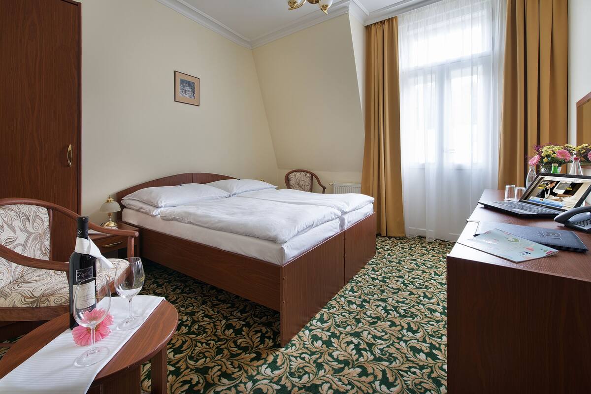 A hotel room with a bed, a desk with a laptop, a bottle of wine, and a glass on the desk.