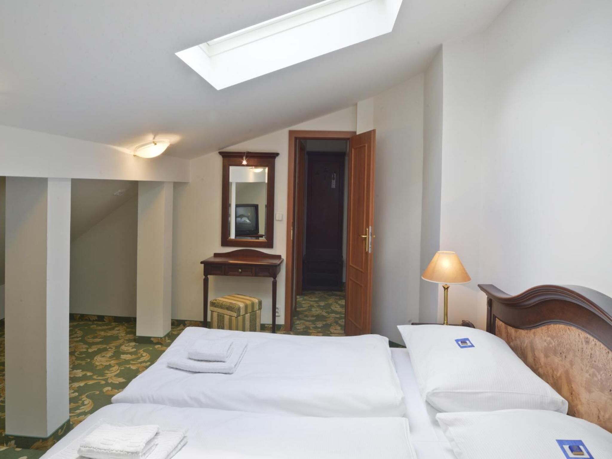 A hotel room with two beds, a desk, a mirror, a lamp, and a skylight.