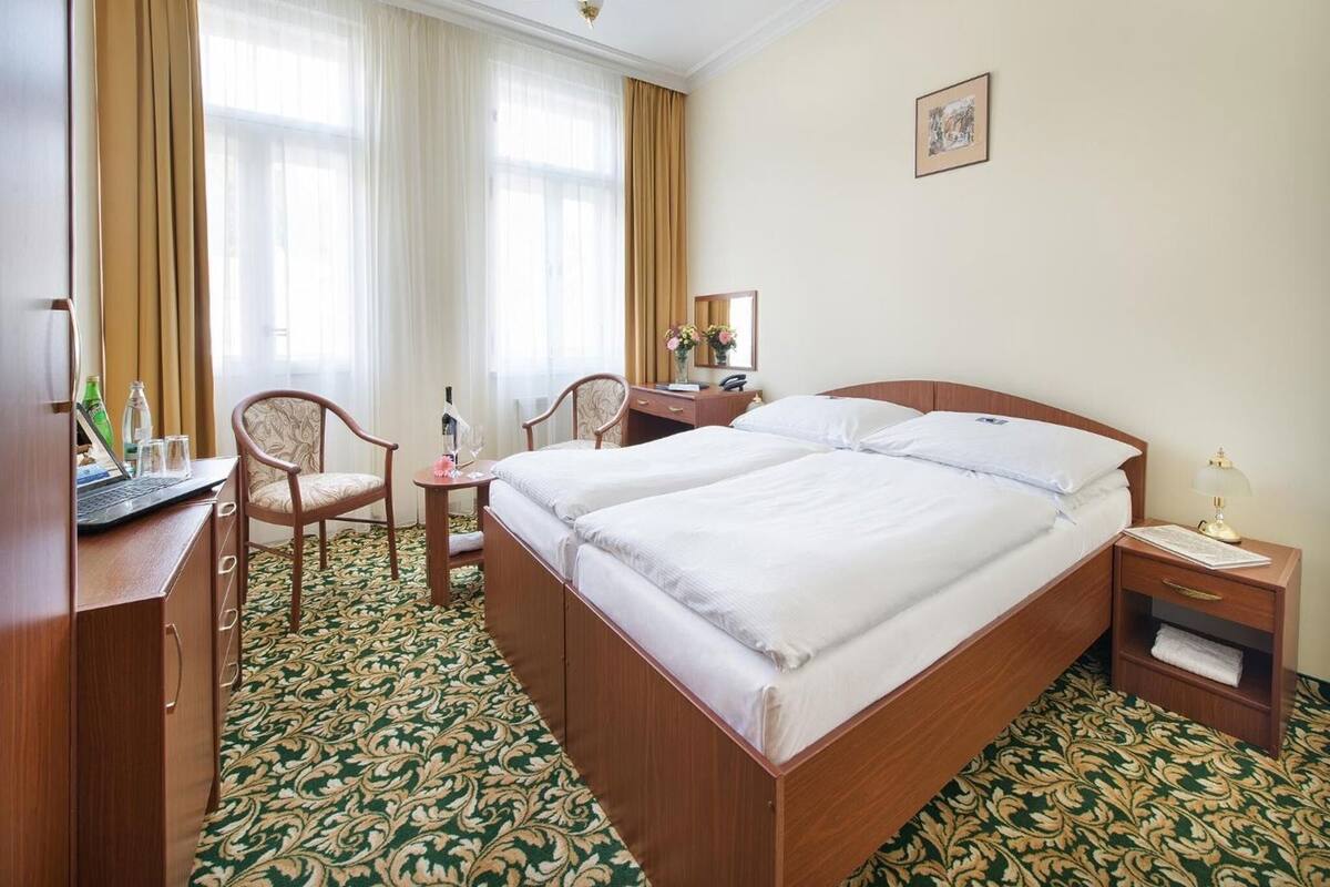 A hotel room with two beds, a desk with a laptop, a chair, a bedside table with a lamp, and a large window with curtains.