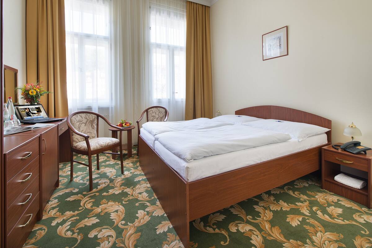 A hotel room with a bed, a desk with a laptop, two chairs, a bedside table with a lamp, and a large window with curtains.