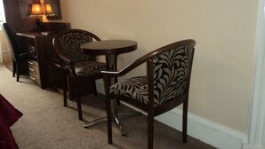 A small, cozy room with a round table and two chairs with patterned upholstery. A desk with drawers and a lamp is visible in the background.