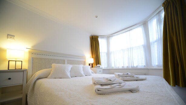 A neatly made bed with white linens and towels, a bedside lamp, and a window with curtains. - Marine View (Brighton)