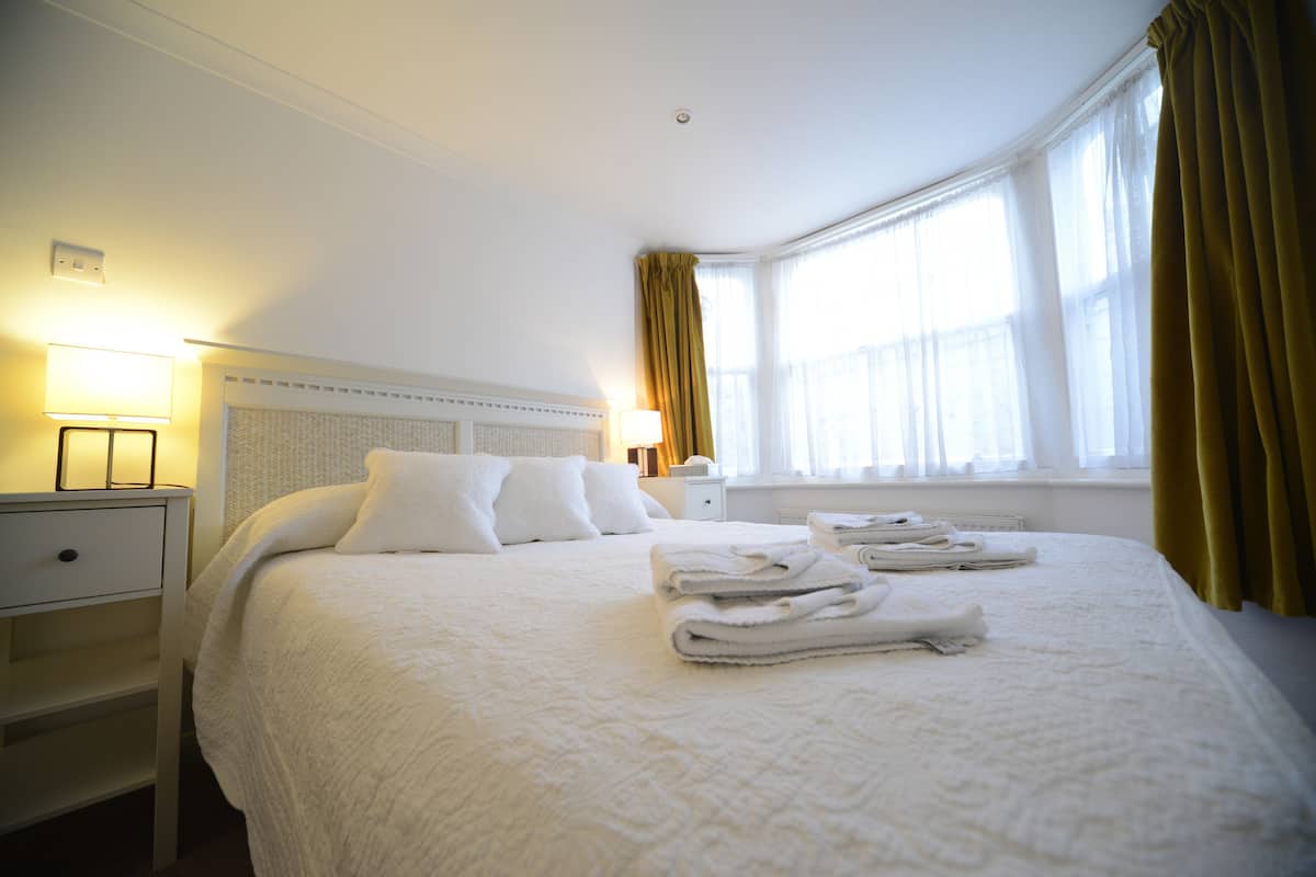 A neatly made bed with white linens and towels, a bedside lamp, and a window with curtains.
