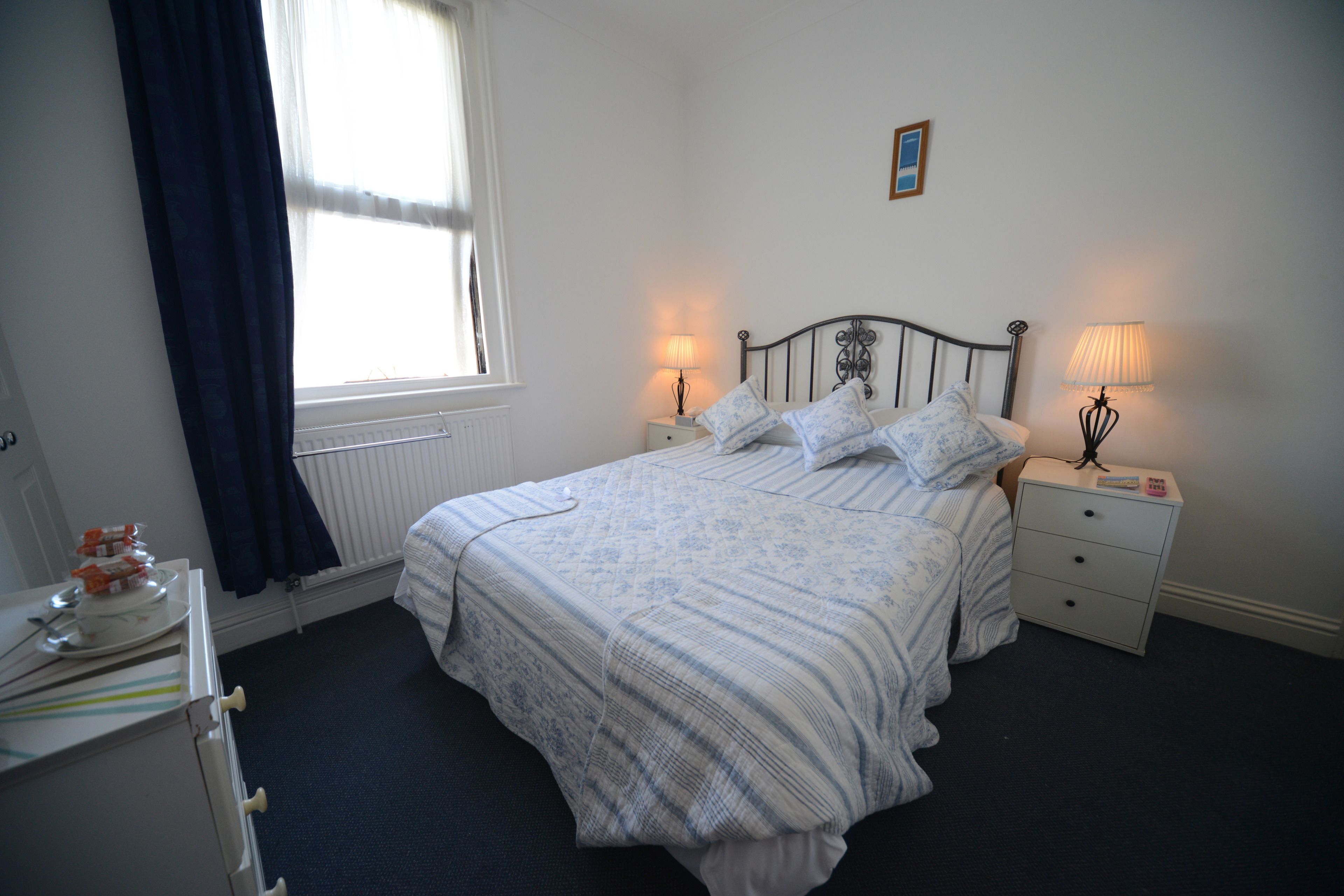 A neatly made bed with a striped bedspread, two bedside tables with lamps, a window with curtains, and a radiator.