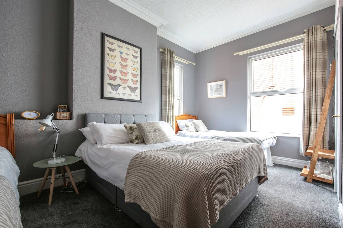 A bedroom with two beds, a bedside table with a lamp, a framed butterfly artwork, and a window with curtains.