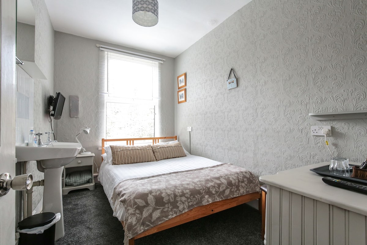 A bedroom with a bed, a bedside table, a lamp, a window, and a wall-mounted clock.