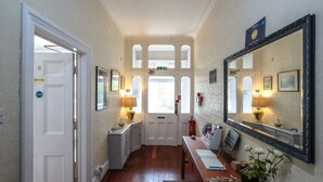 Un pasillo con una puerta blanca marcada “PRIVATE”, un banco con un jarrón de flores y un espejo grande en la pared.