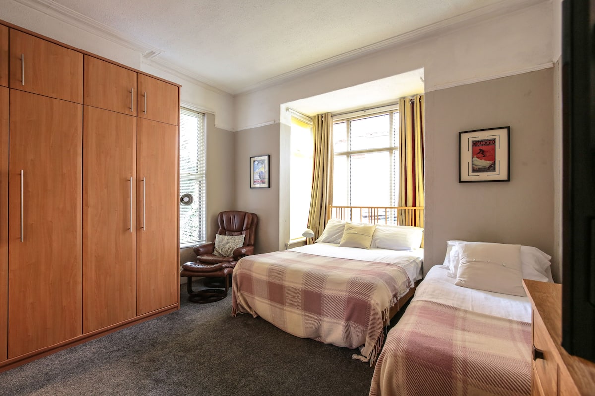 A bedroom with a large wardrobe, a chair, two beds, and a window with curtains.