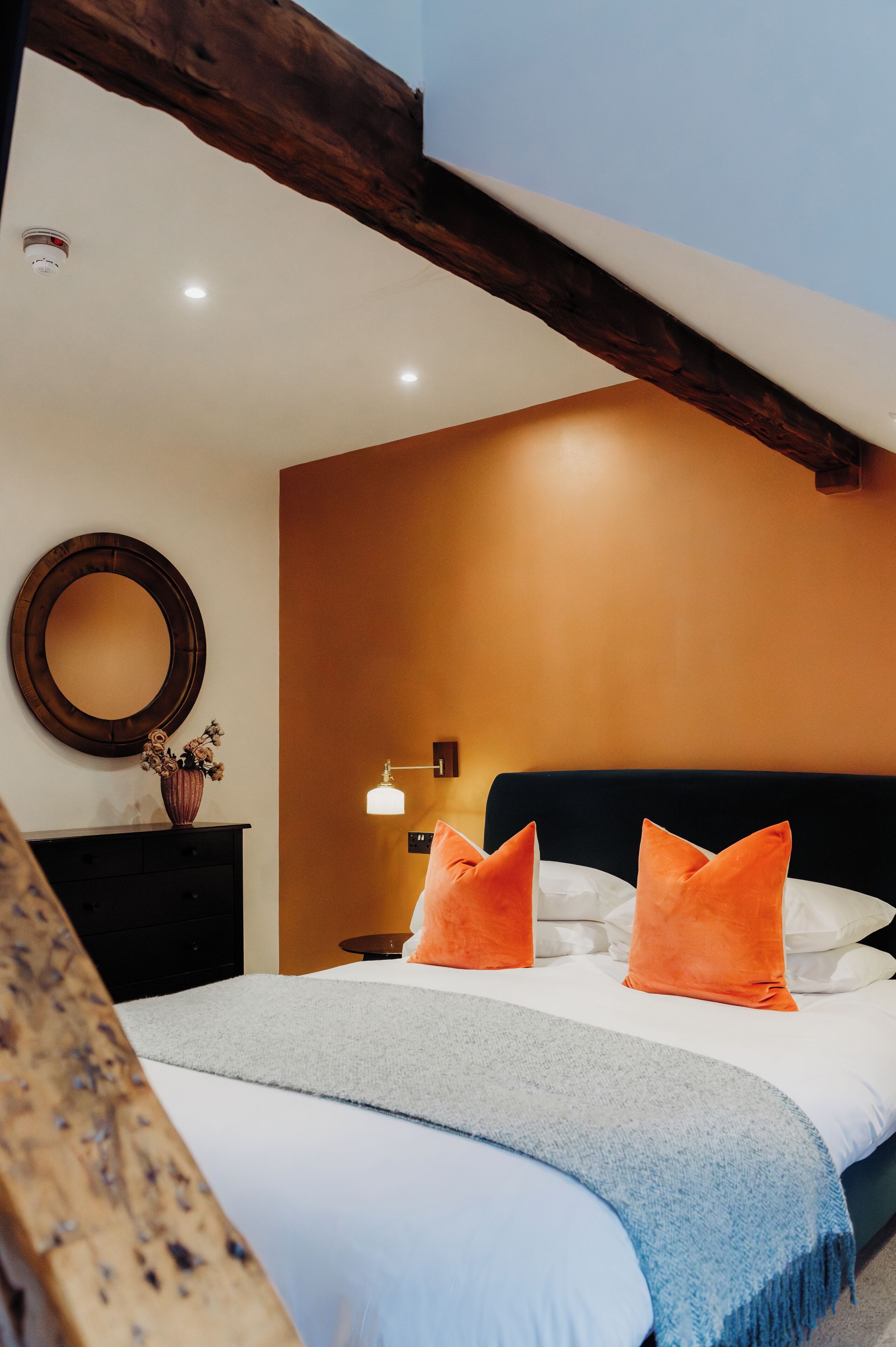 A bedroom with a bed, orange pillows, a bedside table with a vase, and a wall-mounted mirror.
