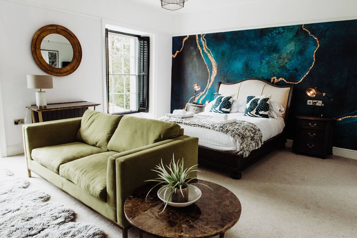 A bedroom with a bed, a green sofa, a round coffee table with a plant, a large mirror, and a dark blue accent wall.