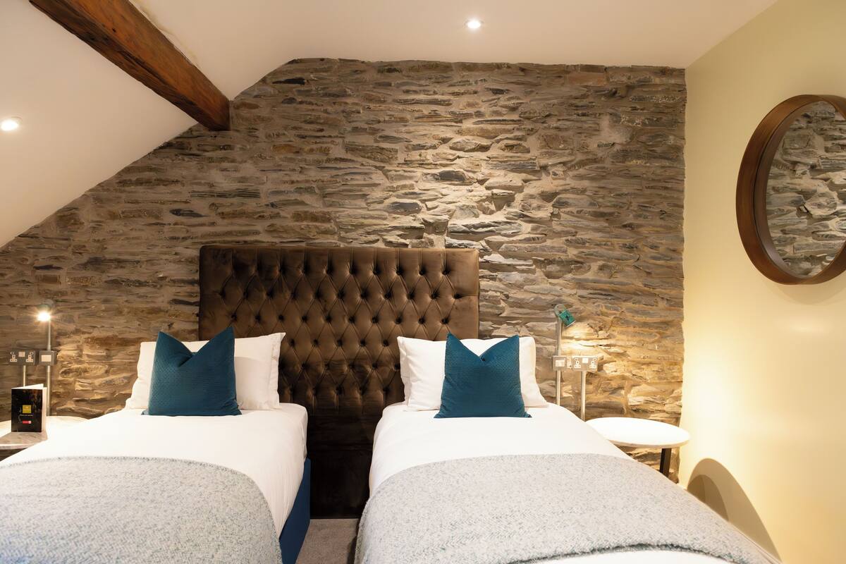 A bedroom with two beds, a stone wall, a tufted headboard, and a round mirror.