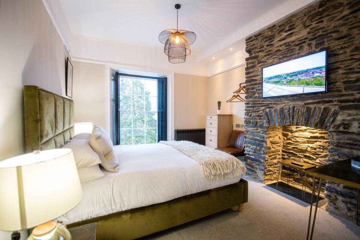 A bedroom with a bed, a lamp, a TV mounted on a stone wall, and a fireplace.