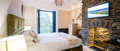 A bedroom with a bed, a lamp, a TV mounted on a stone wall, and a fireplace.