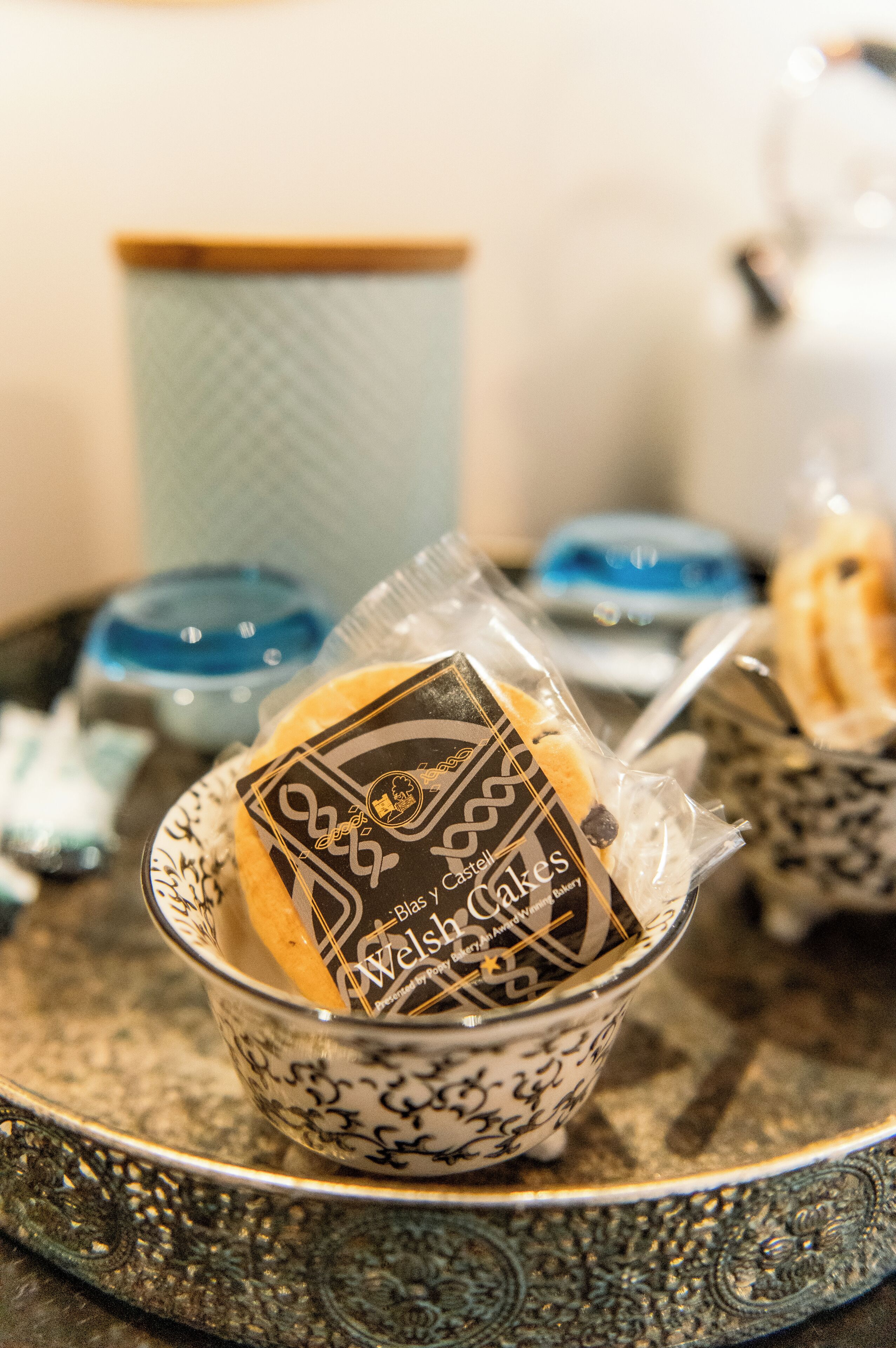 A tray with a decorative bowl containing a packaged item, a spoon, and a blue container in the background.