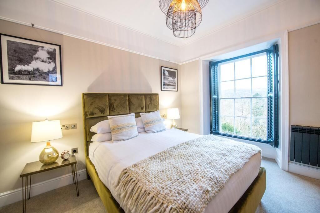 Un dormitorio ordenado con una cama grande, dos mesitas de noche con lámparas, un cuadro enmarcado en la pared y una ventana con cortinas.