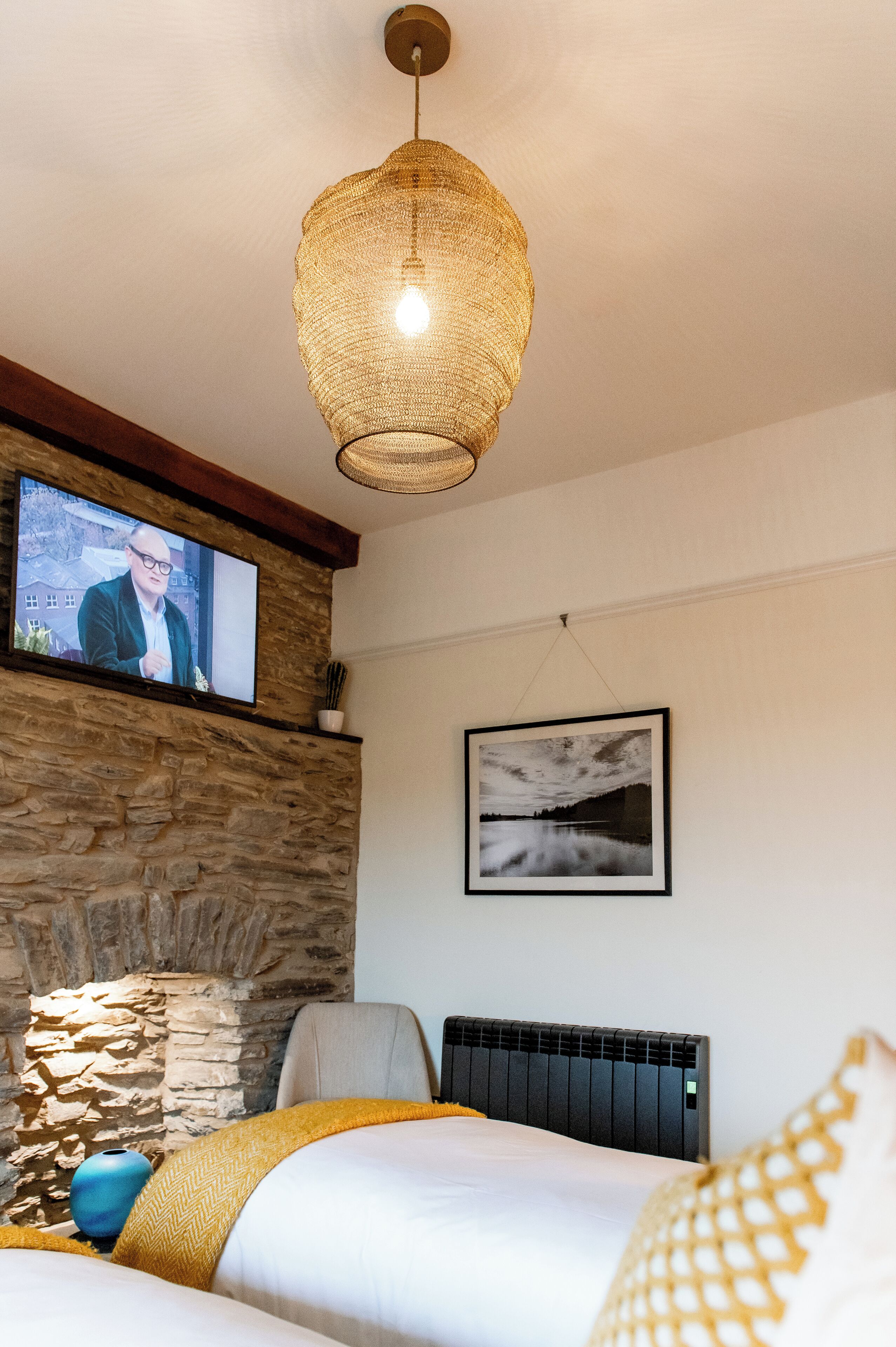 Une chambre avec un mur en pierre, un téléviseur fixé au-dessus, un tableau encadré accroché au mur, un lit recouvert d’une couverture jaune et un radiateur.