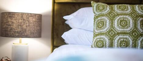 Una cama bien hecha con sábanas blancas y un cojín estampado en verde, una lámpara de noche y un despertador clásico.