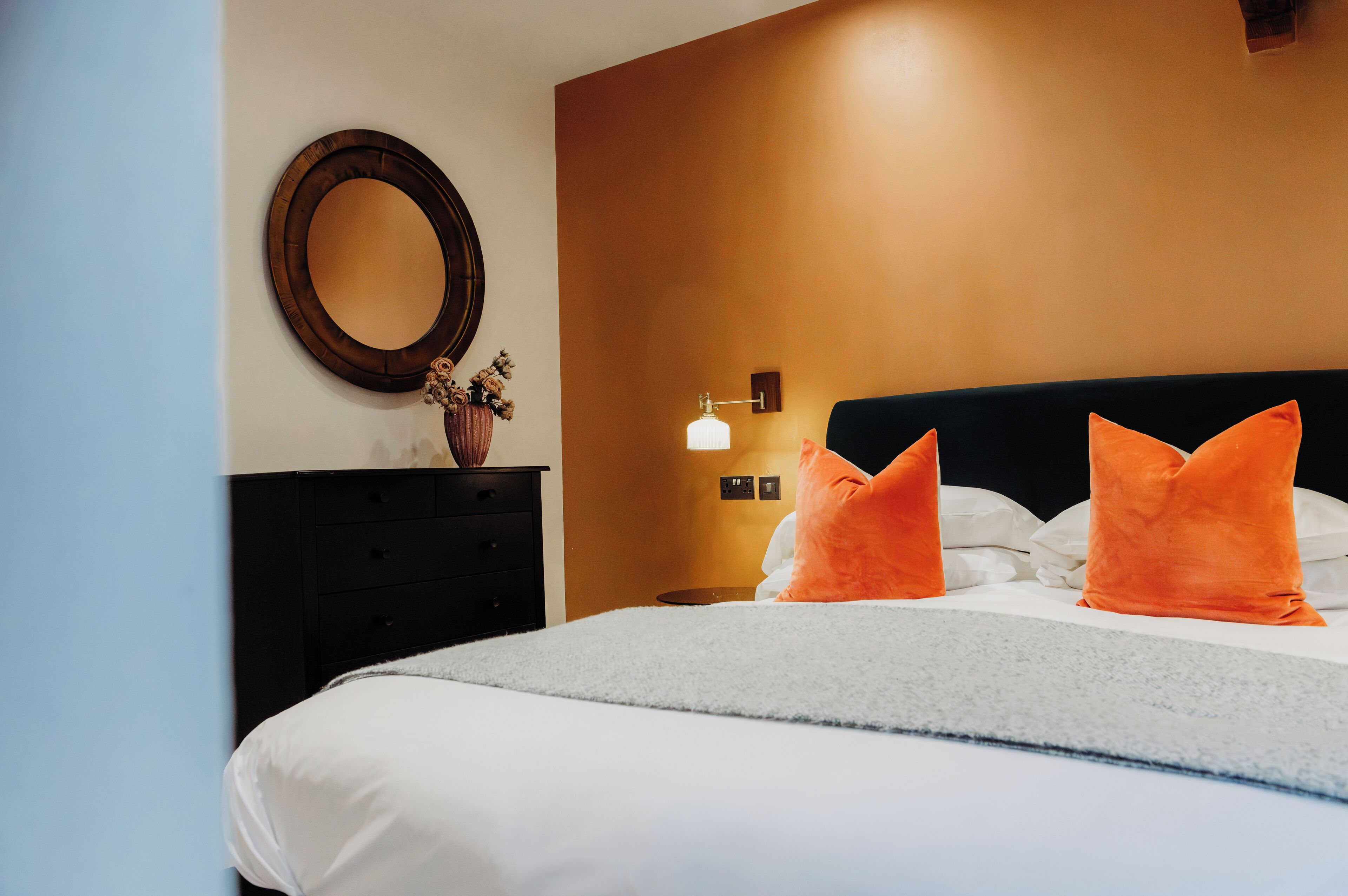 A neatly made bed with white and orange pillows, a round mirror on the wall, and a small table with a vase of flowers.