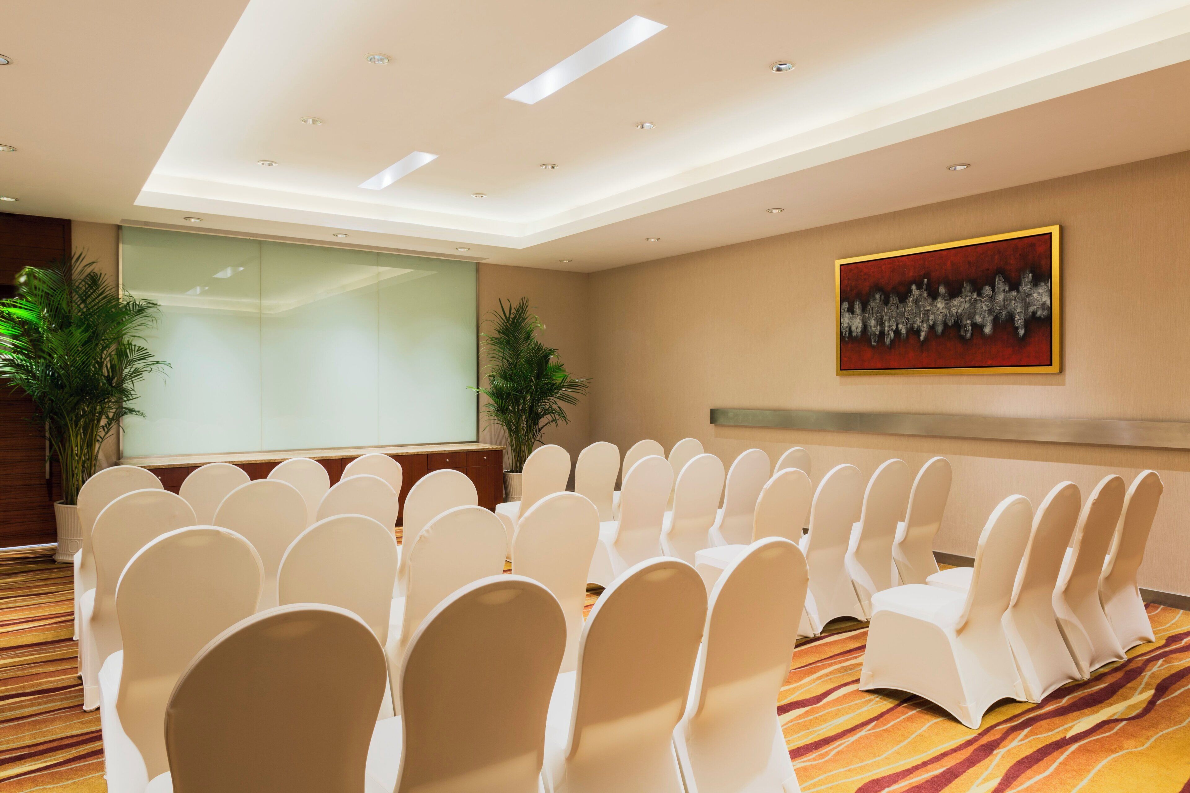 A conference room with rows of white chairs, a projector screen, and a painting on the wall.