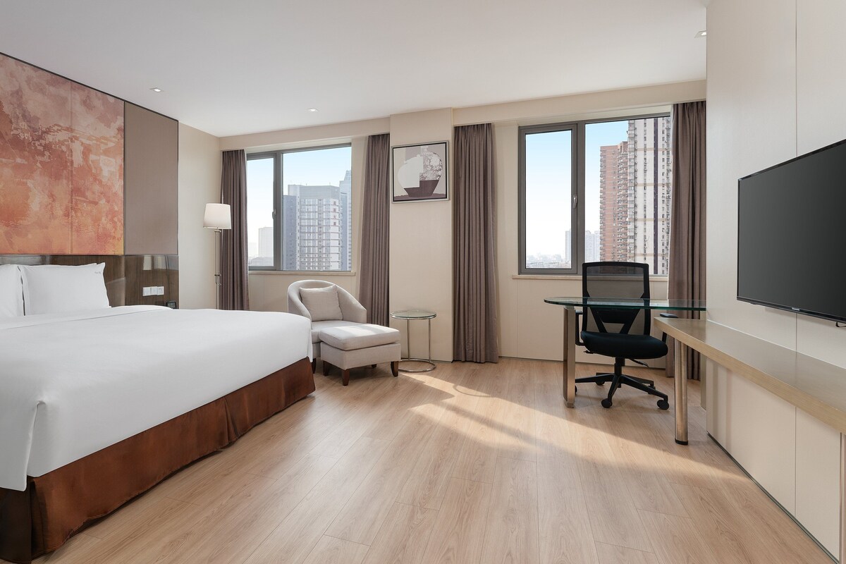 A hotel room with a large bed, a desk with a chair, a TV, and a window with a view of buildings.