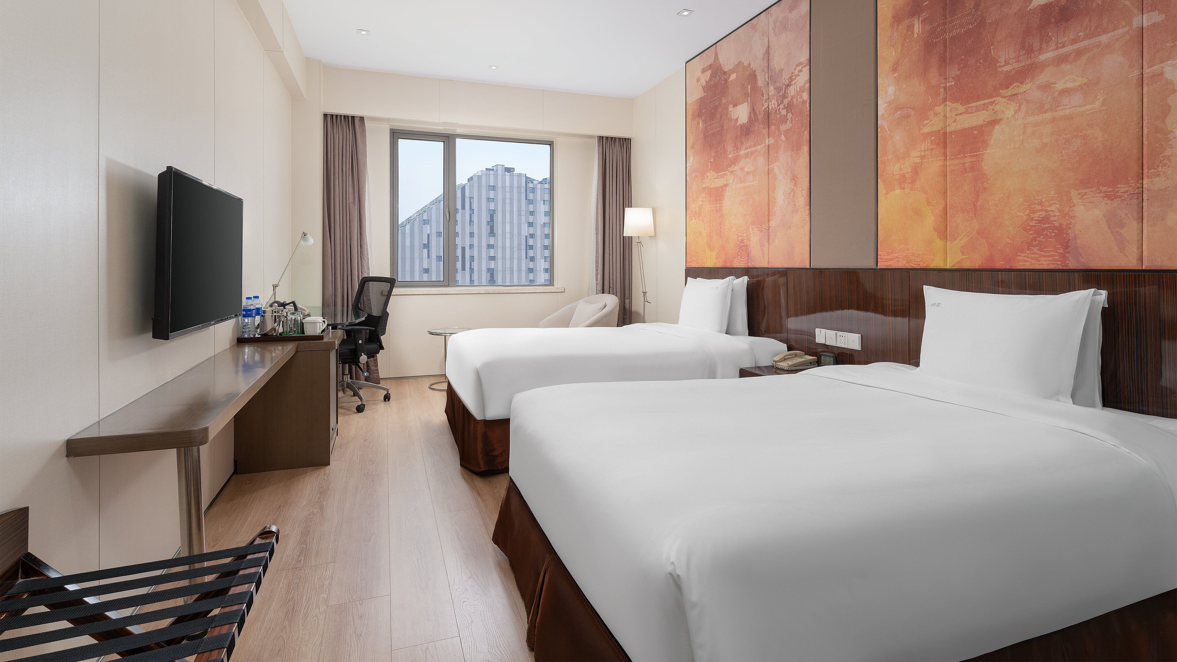 Habitación de hotel con dos camas, un escritorio con televisor, una silla, una lámpara y vistas a edificios.