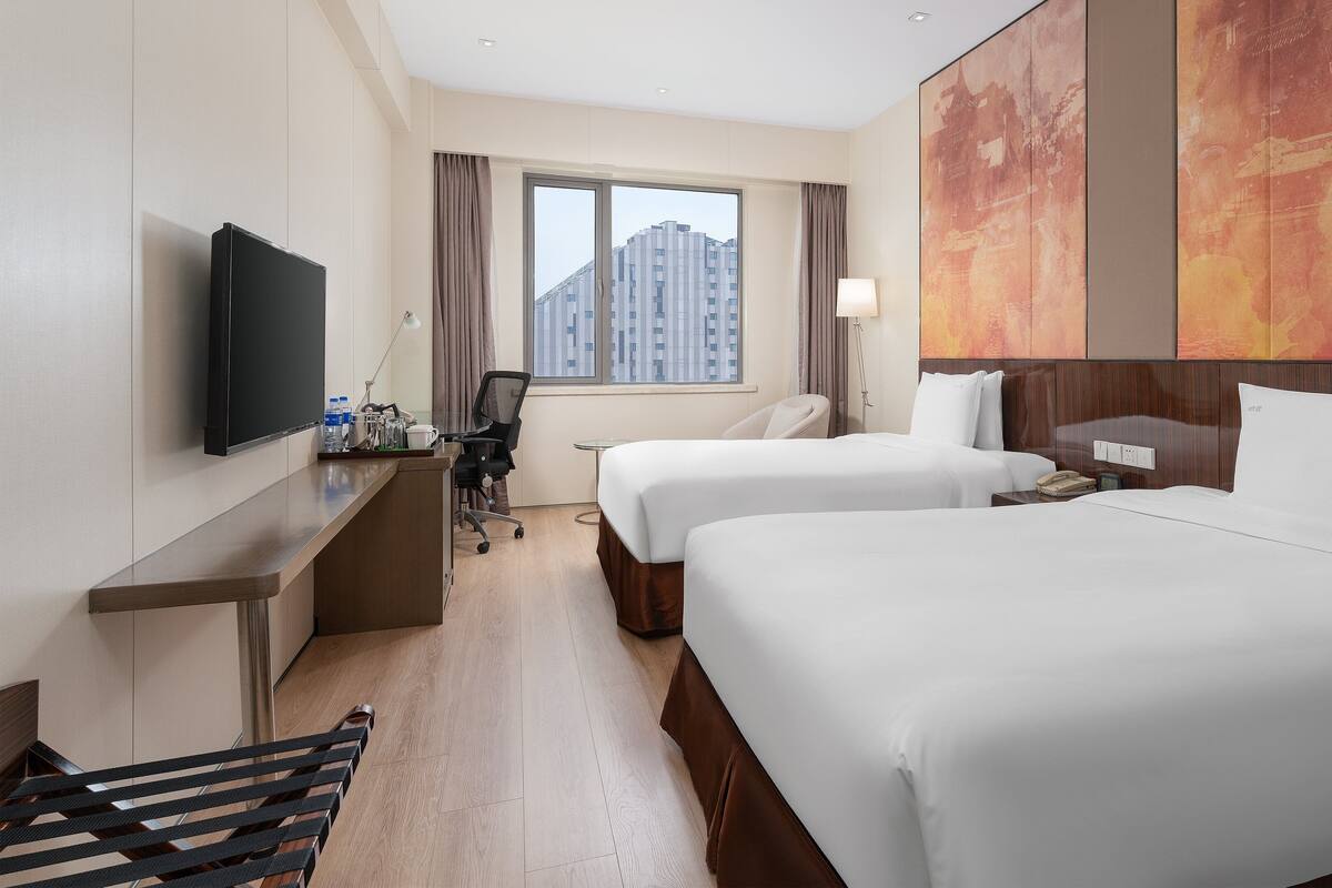 A hotel room with two beds, a desk with a TV, a chair, a lamp, and a view of buildings.