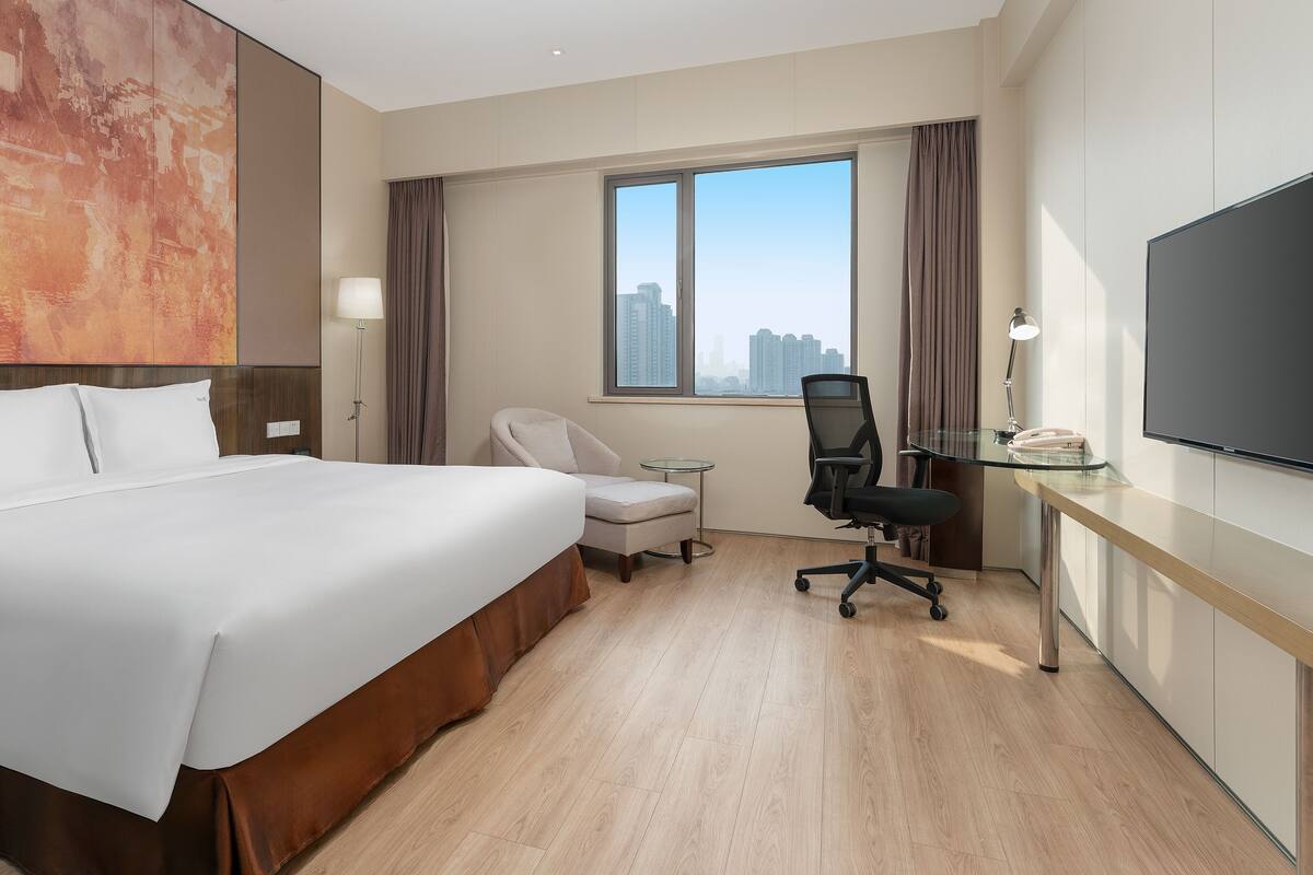 A hotel room with a large bed, a desk, a chair, a TV, and a window with a city view.