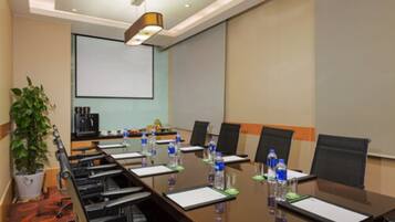 Un salón de conferencias con una larga mesa, sillas, botellas de agua y una pantalla de proyector.