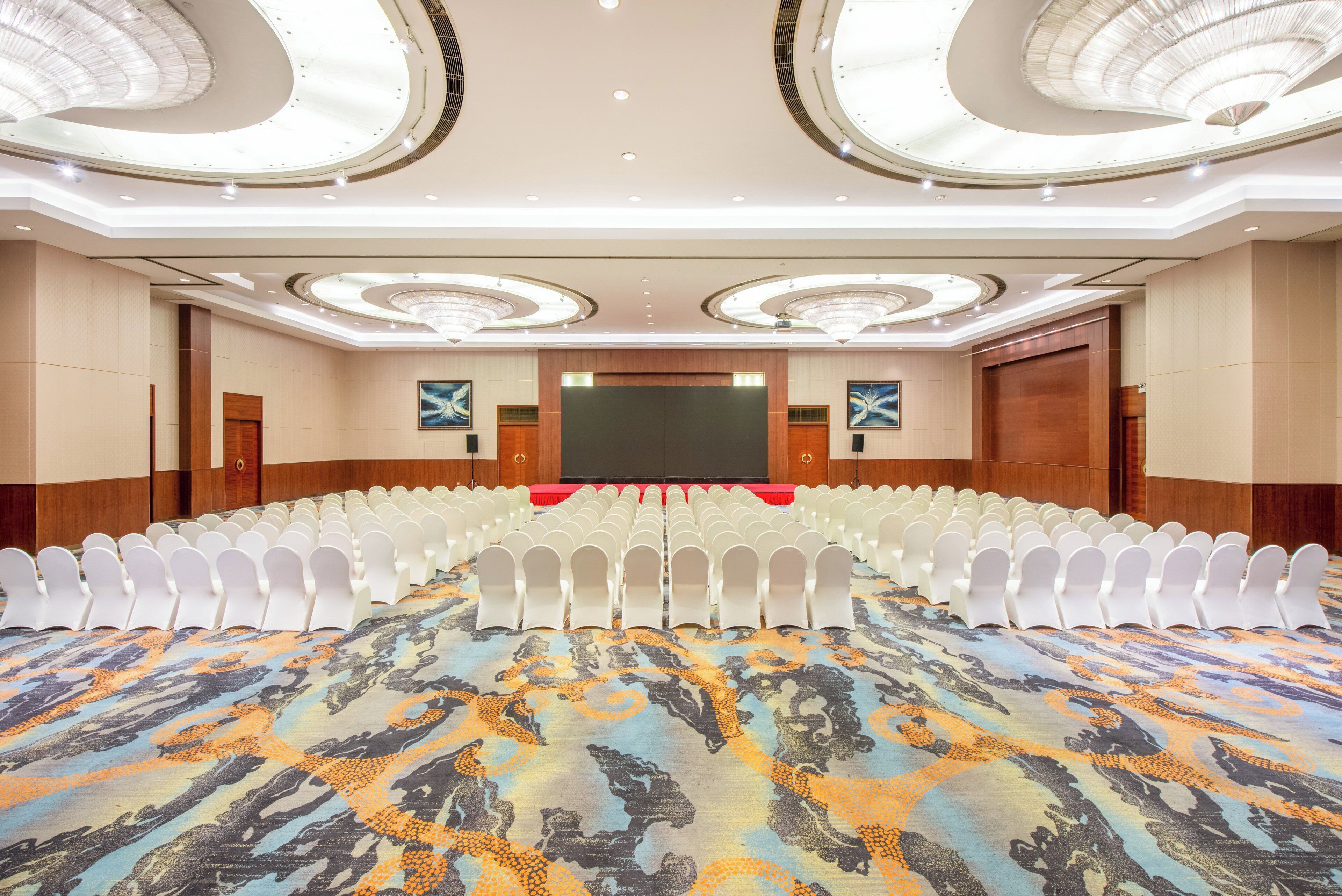 A conference room with rows of white chairs, a large screen, and a patterned carpet.
