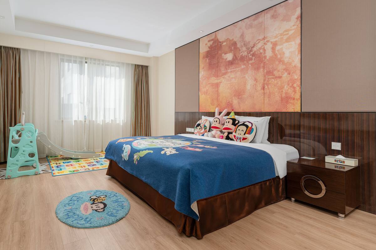 A hotel room with a bed, a television, a desk, a desk lamp, a chair, a window with curtains, and a large abstract painting on the wall.