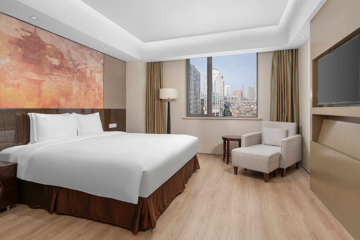 A hotel room with a large bed, a chair, a TV, a window with a city view, and a large abstract painting on the wall.