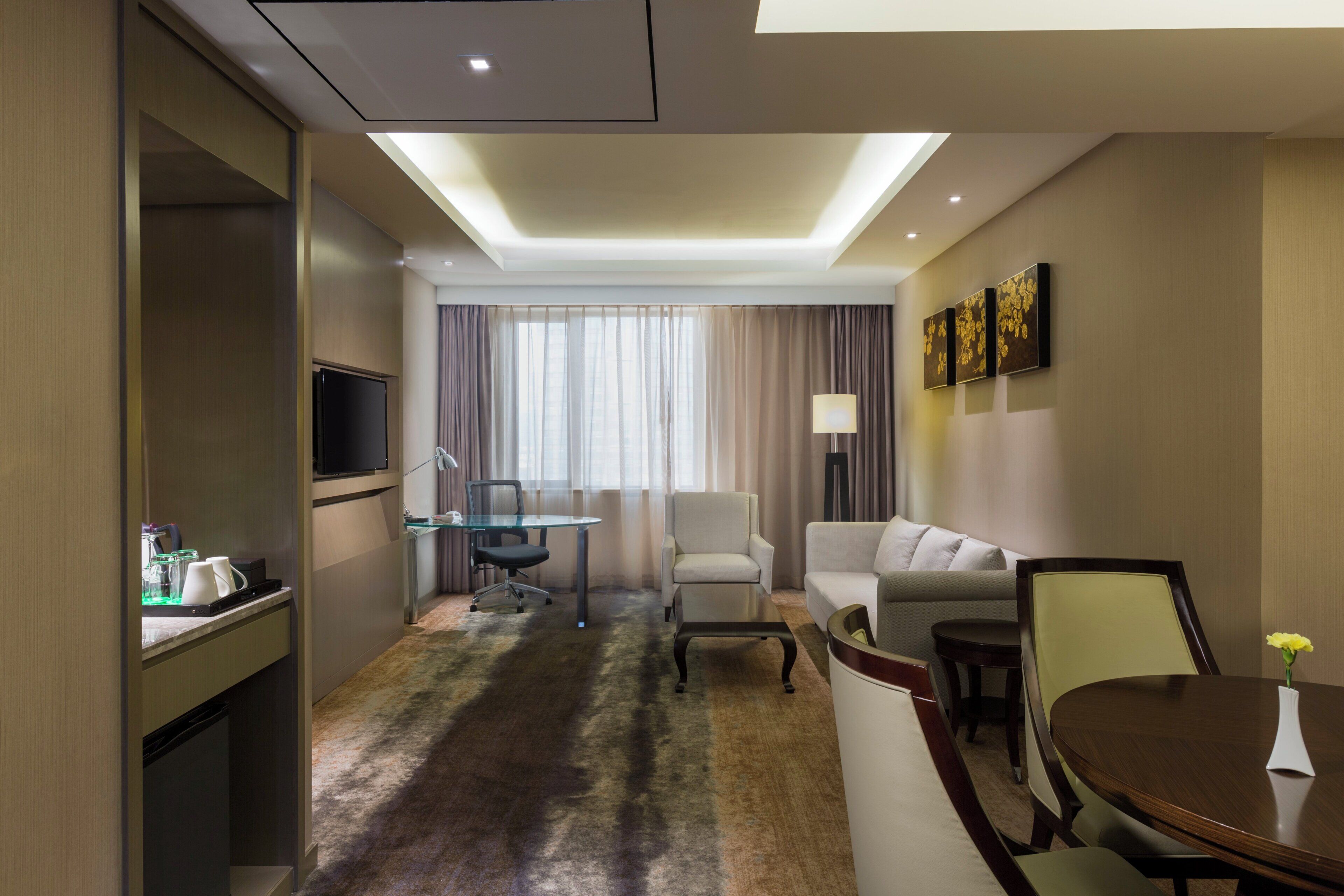 Habitación de hotel con televisión de pantalla plana, mesa de comedor de vidrio, sofá, mesa de centro y lámpara.