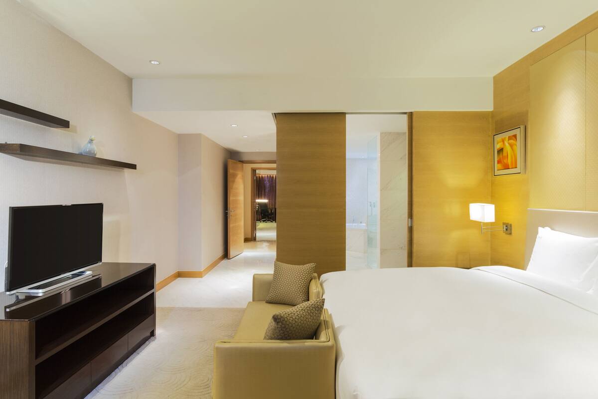 A hotel room with a bed, a TV, a sofa, and a hallway leading to another room.