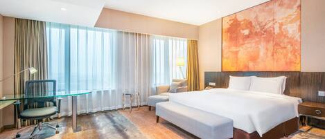 Habitación de hotel con una cama grande, un escritorio con una silla, un sofá y un ventanal con cortinas.