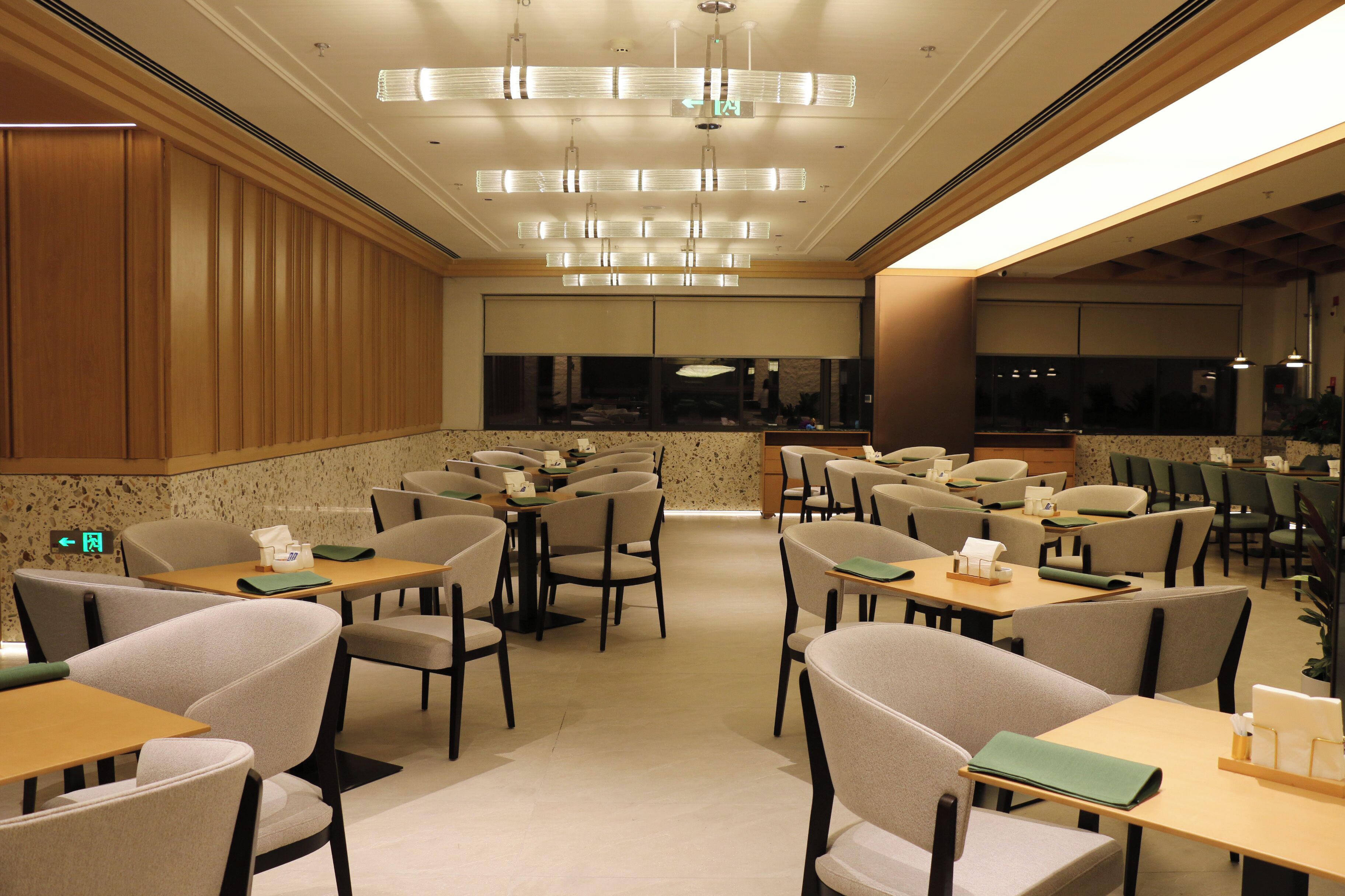 A modern restaurant with multiple tables and chairs, a central chandelier, and a view of the cityscape through large windows.